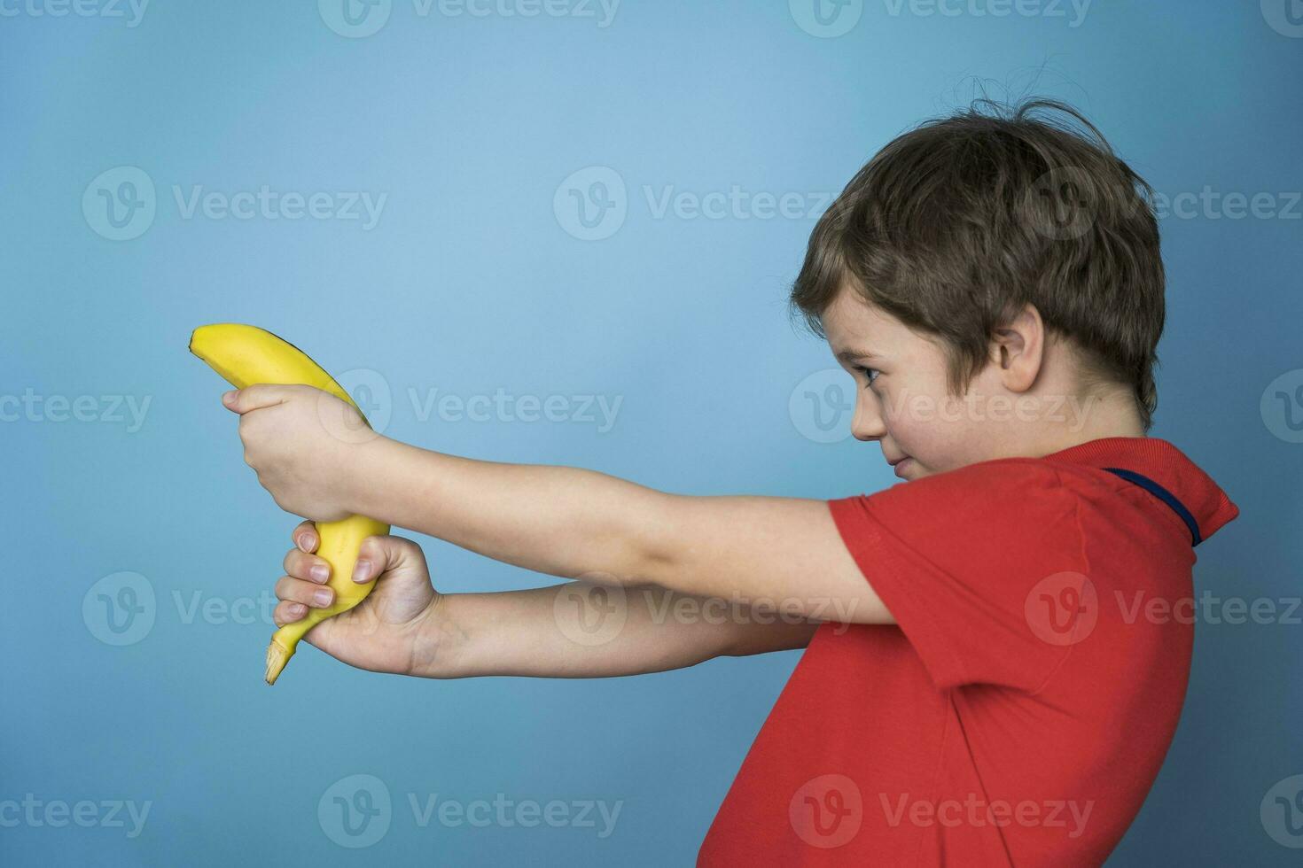 a cute Caucasian boy in a red T-shirt aggressively and cheerfully shoots a banana like a pistol. photo