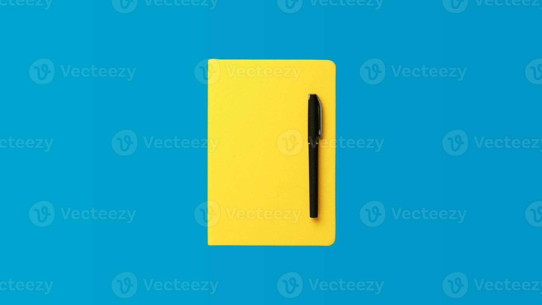 Photo of closed yellow notebook and ball-point pen over blue background. Minimalist image of closed diary and black pen as back to school concept.