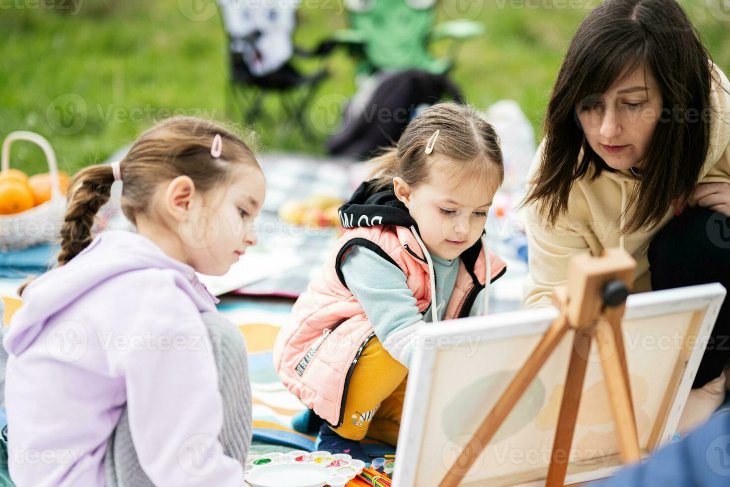 contento joven familia, madre y hijas teniendo divertido y disfrutando al aire libre en picnic cobija pintura a jardín primavera parque, relajación. foto