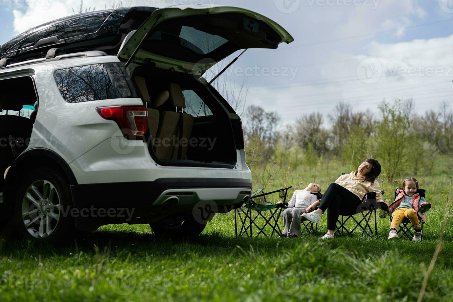 Mother with two daughters sit on chair against car open trunk on picnic. photo