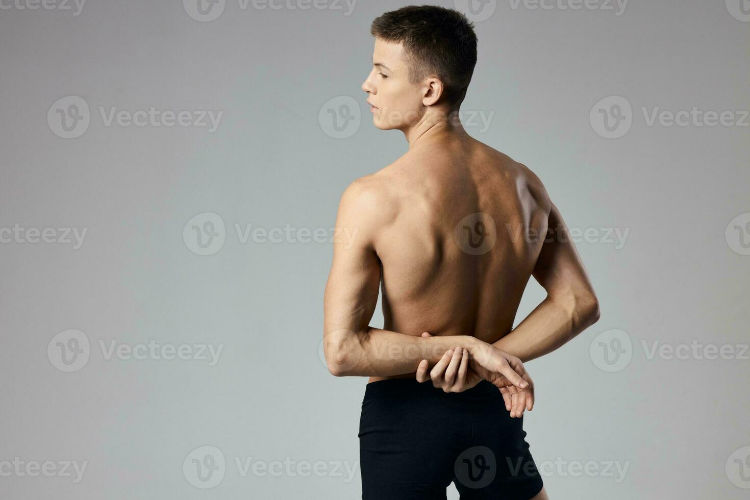 un hombre con bombeado arriba músculos sostiene su manos detrás su espalda en un gris antecedentes y detrás foto