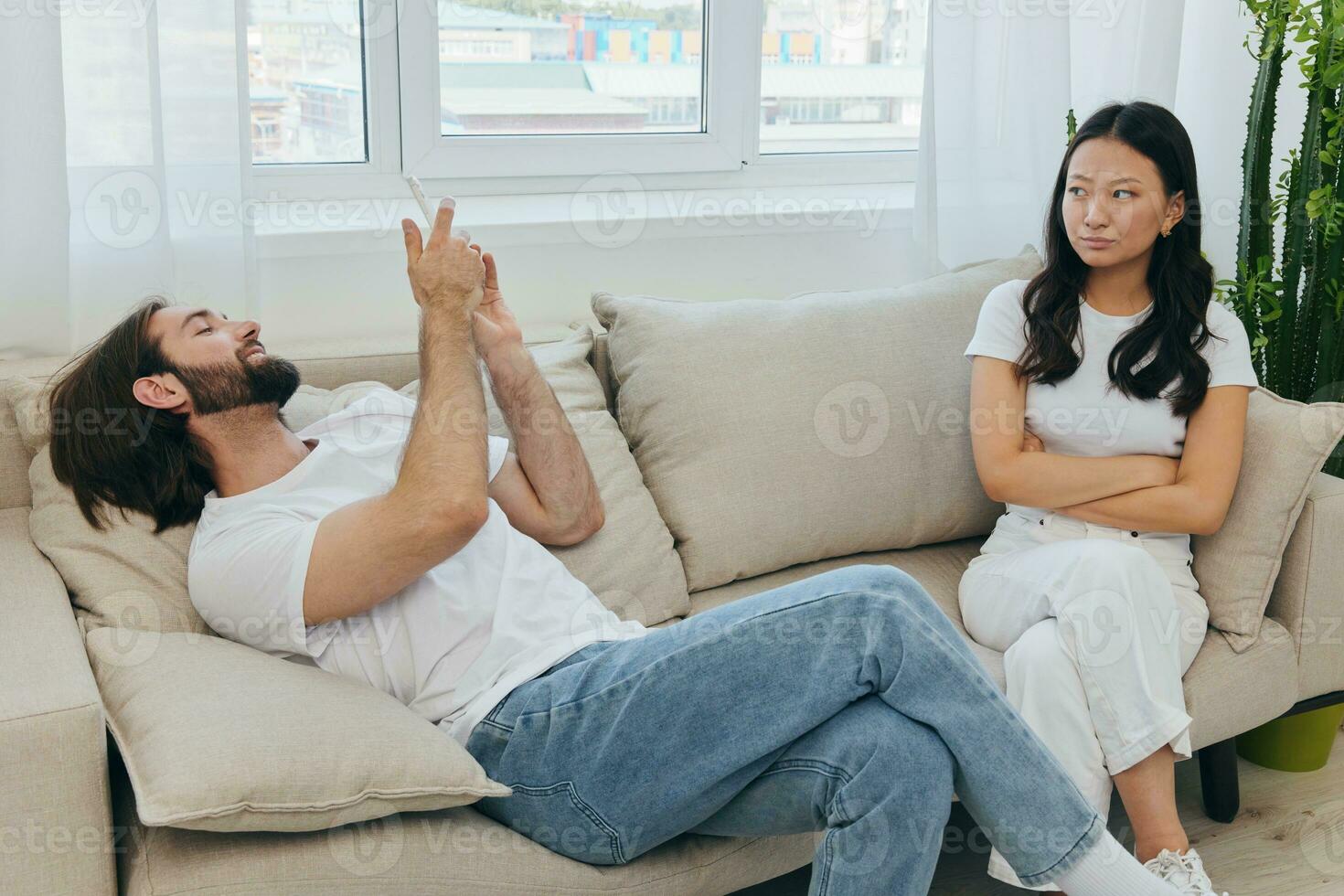 A man looks at his phone screen and smiles during an argument with his girlfriend. The angry and hurt woman looks in his direction and is sad. Family discord at home, phone addiction photo