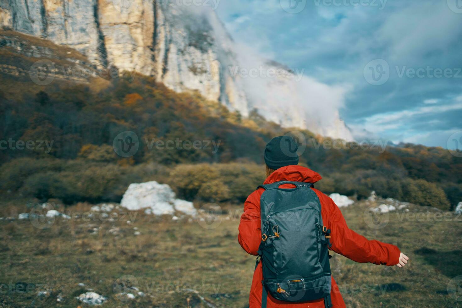 a woman in a red jacket with a backpack and in a warm hat walks on the grass in the fall in the mountains photo