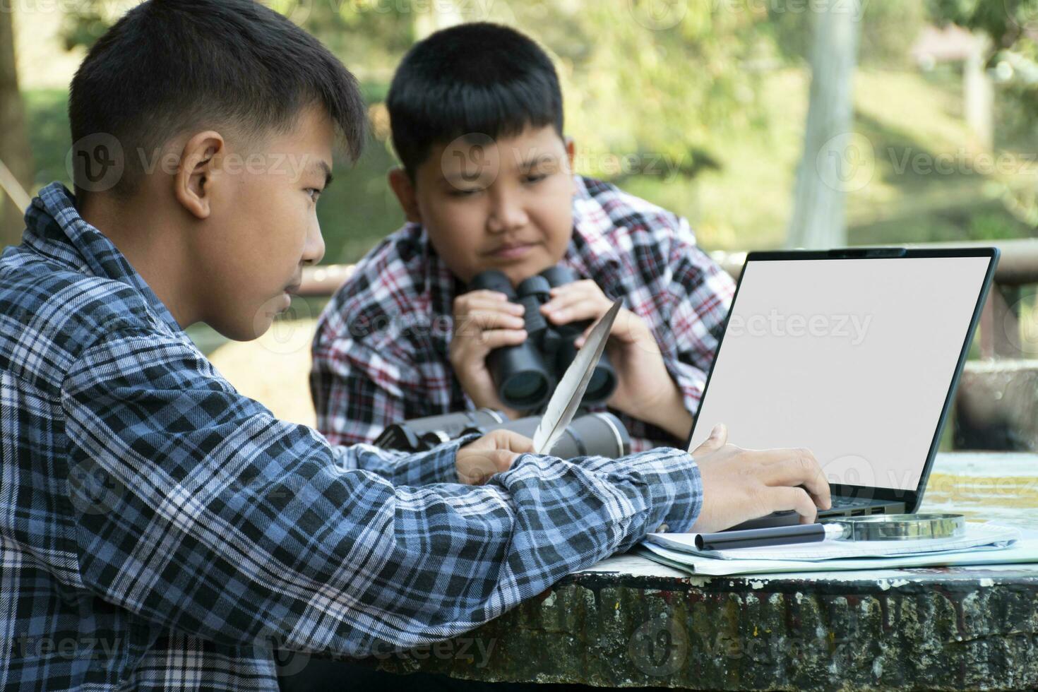 Young asian boy holds bird's feather and using laptop to store and seach birds' information with his friend which holding binoculars behind, selective focus on front boy, nature study concept. photo
