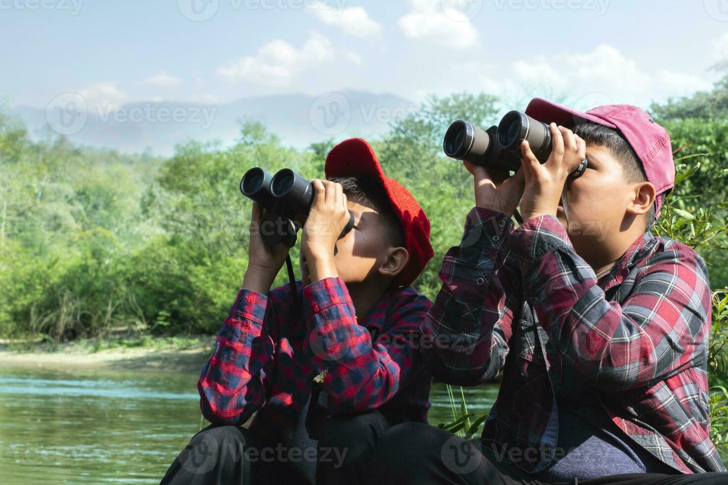 Asian boys sitting, holding binoculars and looking up to watch the birds which flying on sky and sitting on tree branches in local national park to study birds, nature, environment and landscape. photo