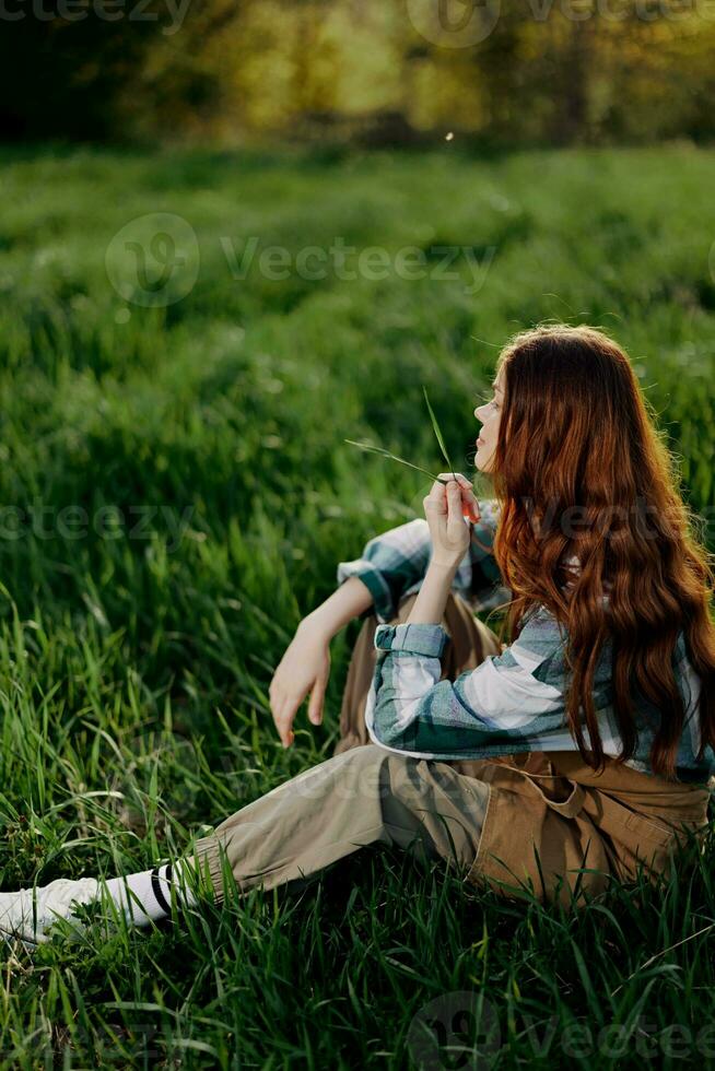 A woman sits on the green grass in a park with her back to the camera and relaxes in nature in the summer sunset evening light photo