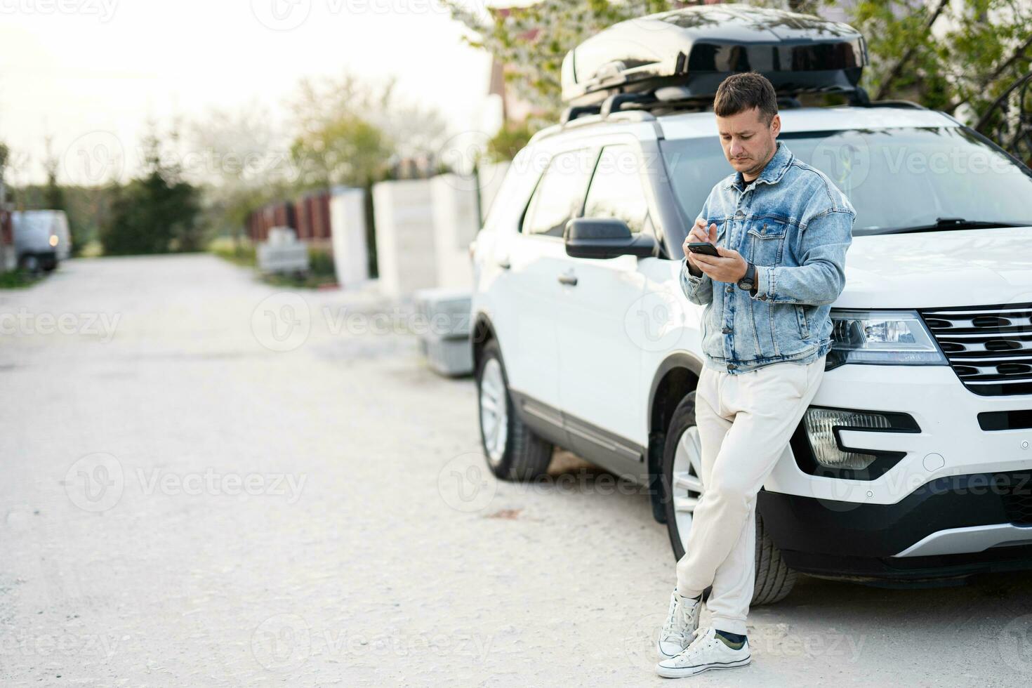 Man in jeans jacket with phone in hand standing near white american suv car with roof rack box. photo