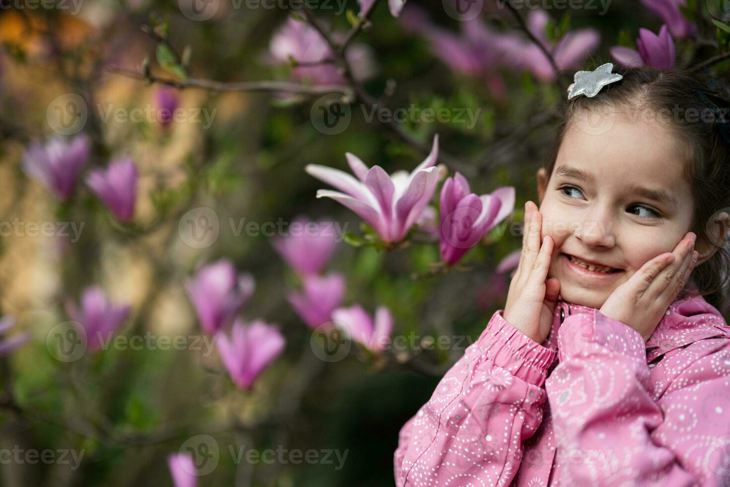 Preschooler girl in pink jacket enjoying nice spring day near magnolia blooming tree. Springtime activities. photo