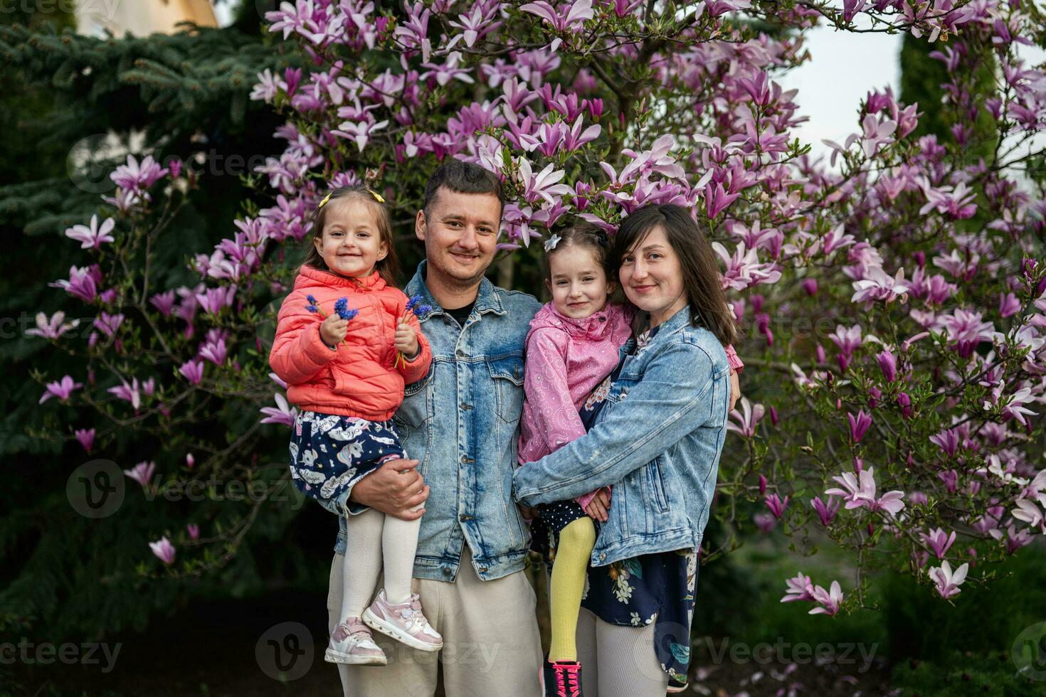 contento familia con dos hijas disfrutando bonito primavera día cerca magnolia floreciente árbol. foto