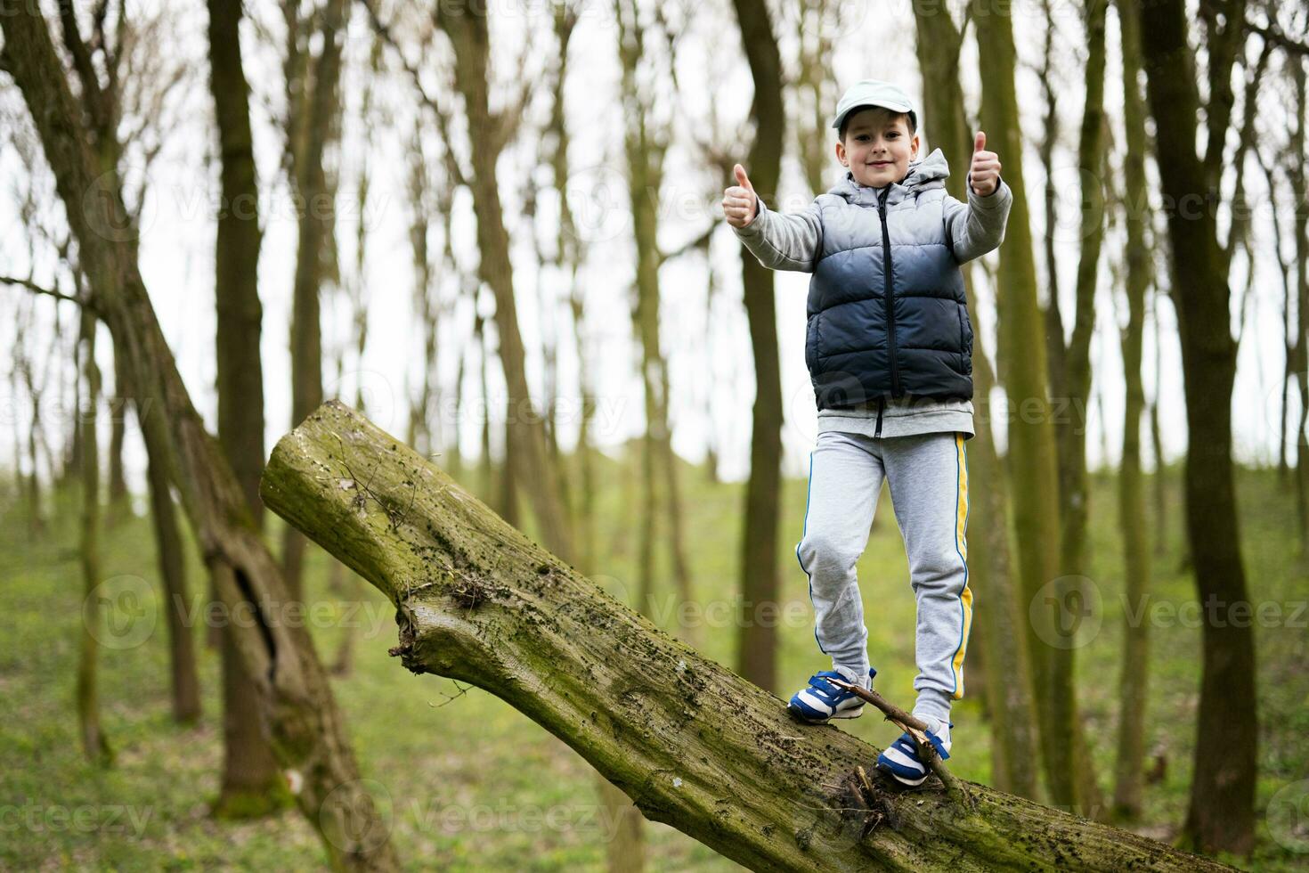 Boy climbed a felled tree in spring forest and show thumbs up. Happy childhood moments. photo