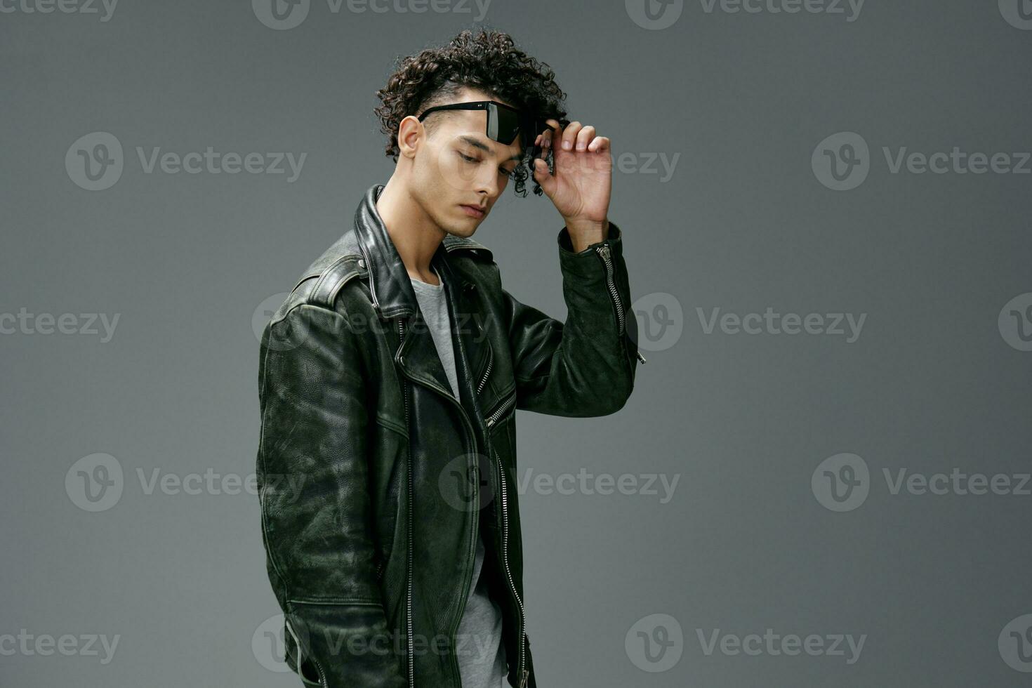 Side of view of stylish tanned curly man leather jacket trendy sunglasses posing isolated on gray studio background. Cool fashion offer. Huge Seasonal Sale New Collection concept. Copy space for ad photo