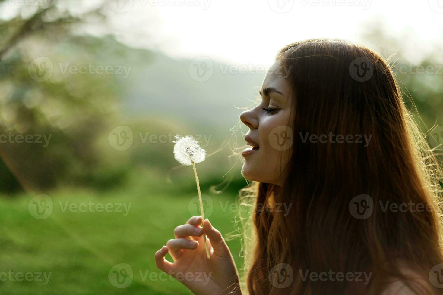 retrato de un joven mujer con un diente de león flor en su mano soplo en eso y sonriente en contra el verde verano césped en el rayos de el ajuste Dom en naturaleza foto