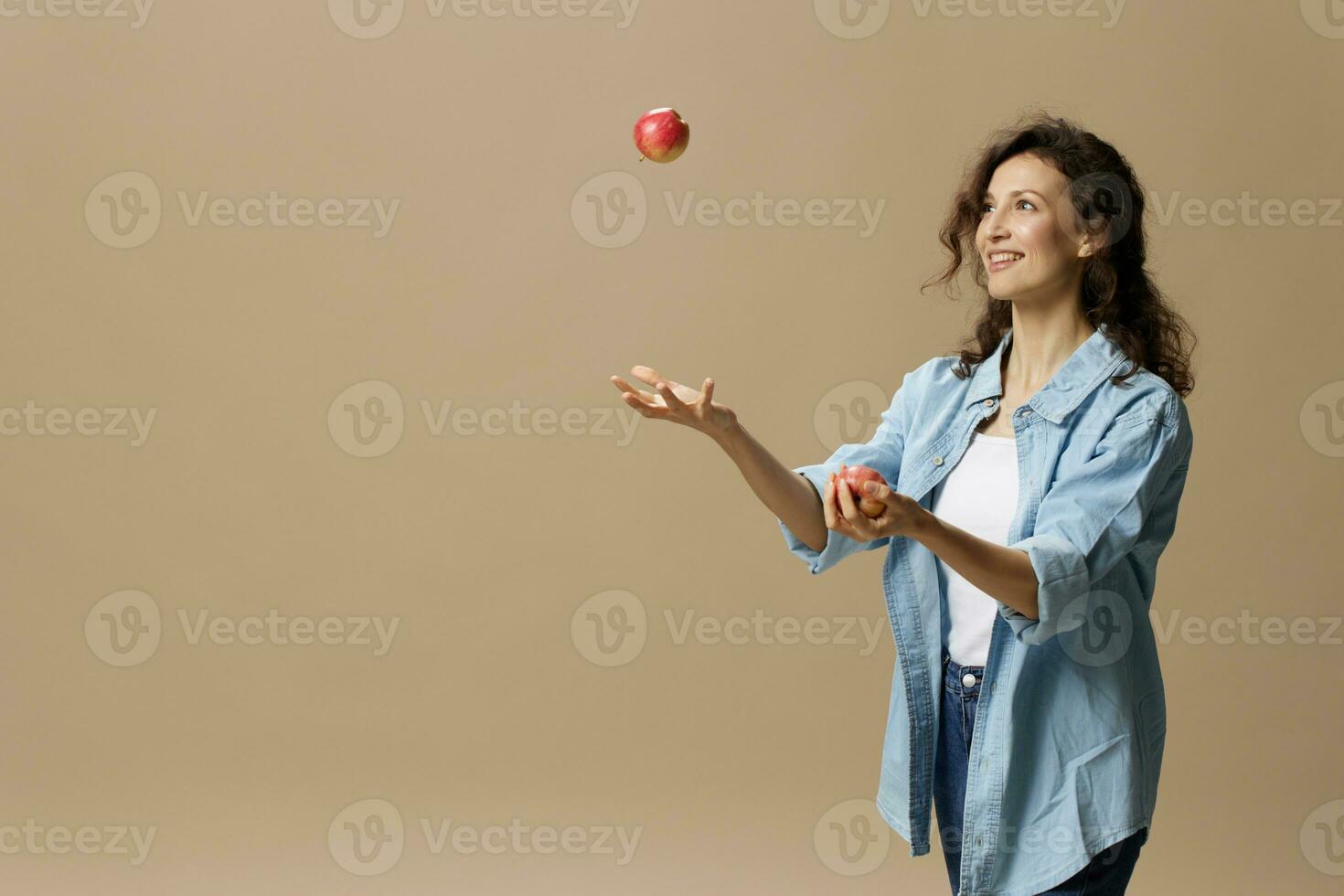 Happy smiling focused curly beautiful female in jeans casual shirt juggling with apple posing isolated on over beige pastel background. Healthy food. Natural eco-friendly products concept. Copy space photo