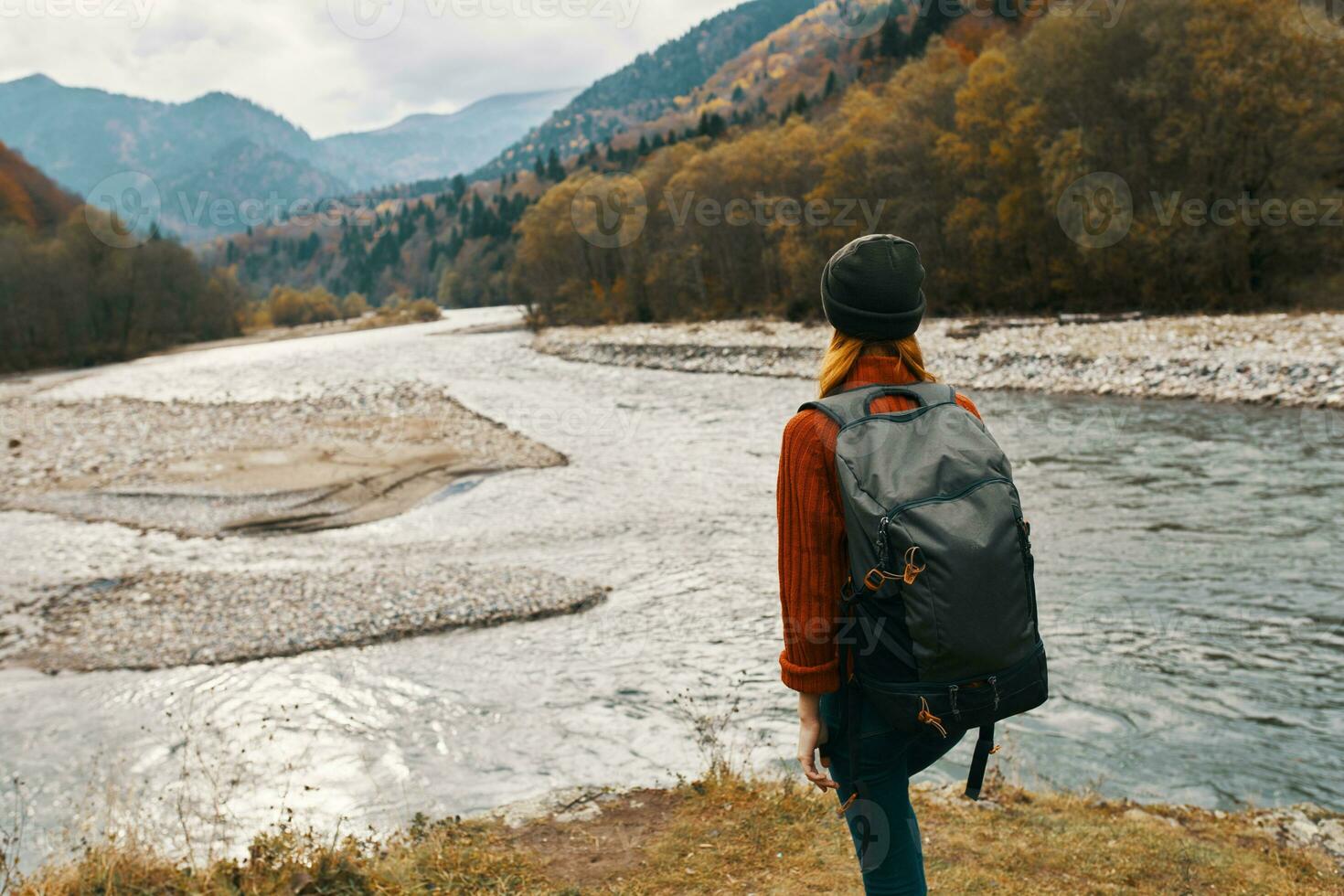 a woman in a red jacket and a backpack on her back are walking on the banks of the river in the mountains photo