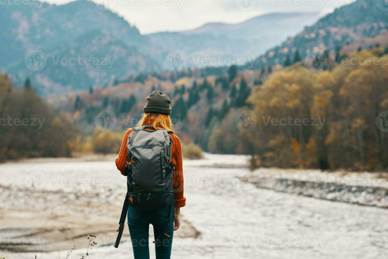 woman in a red sweater cap with a backpack on the river bank and mountains in the distance photo