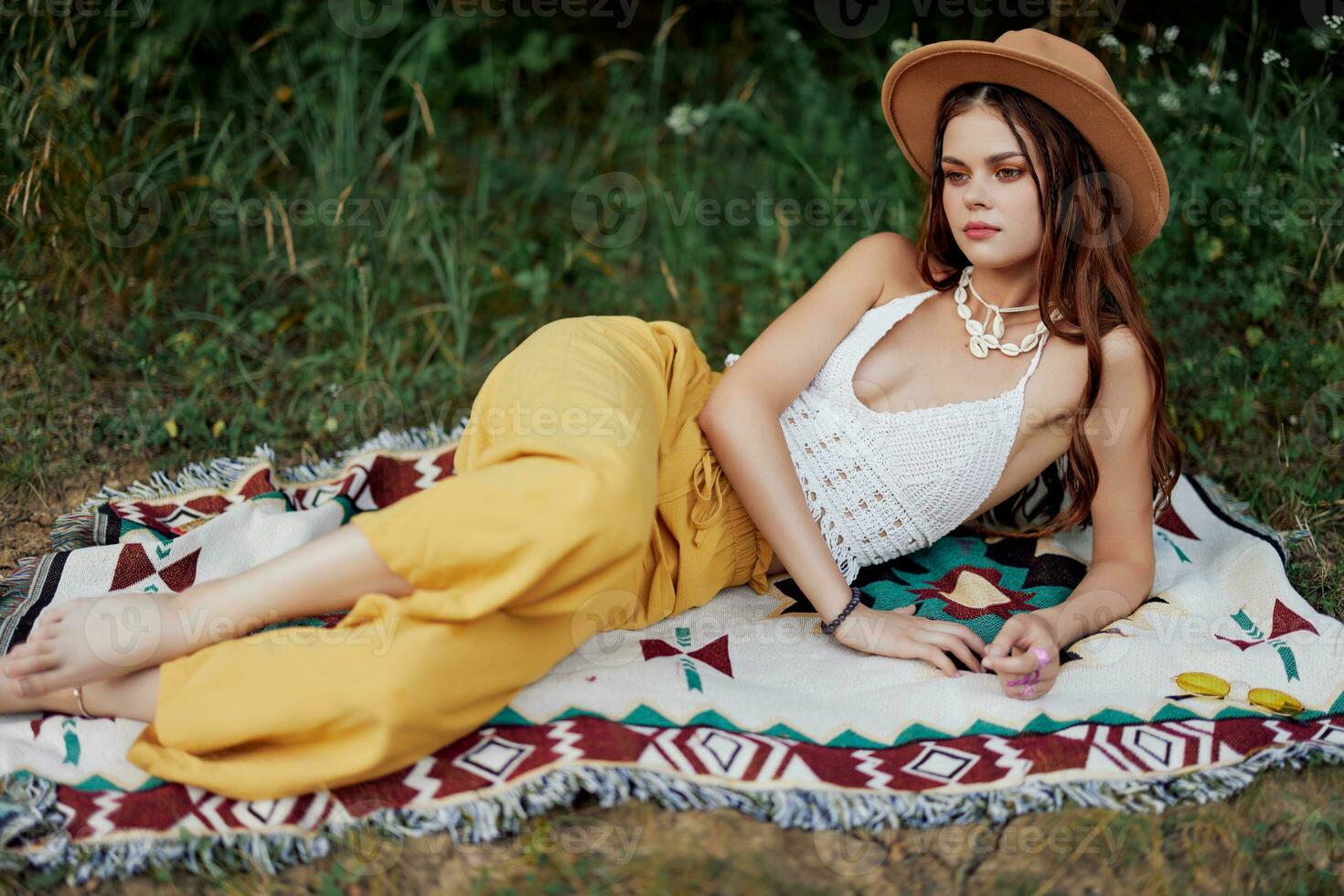 A woman in eco-friendly clothing in a hippie look is lying on a colored plaid smiling and looking at an autumn sunset in nature in the park. Lifestyle on a journey of harmony with the world photo