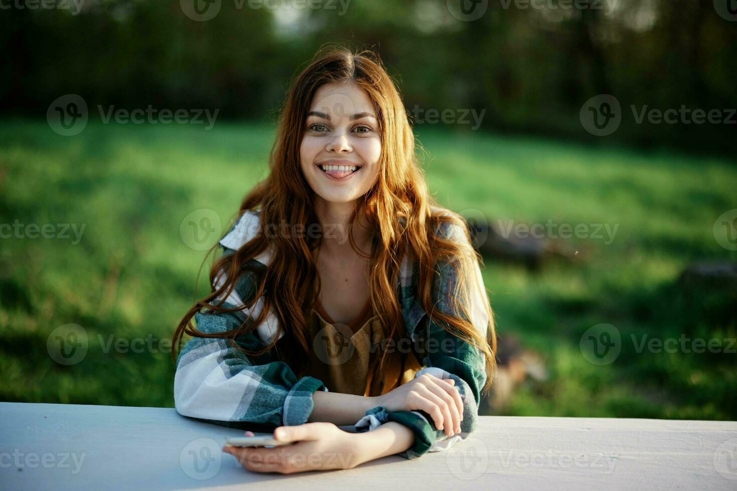 Girl with phone in hand sitting in nature in the park at the table smiling beautifully and looking at the camera with her red hair lit by the sunset sunlight of summer photo