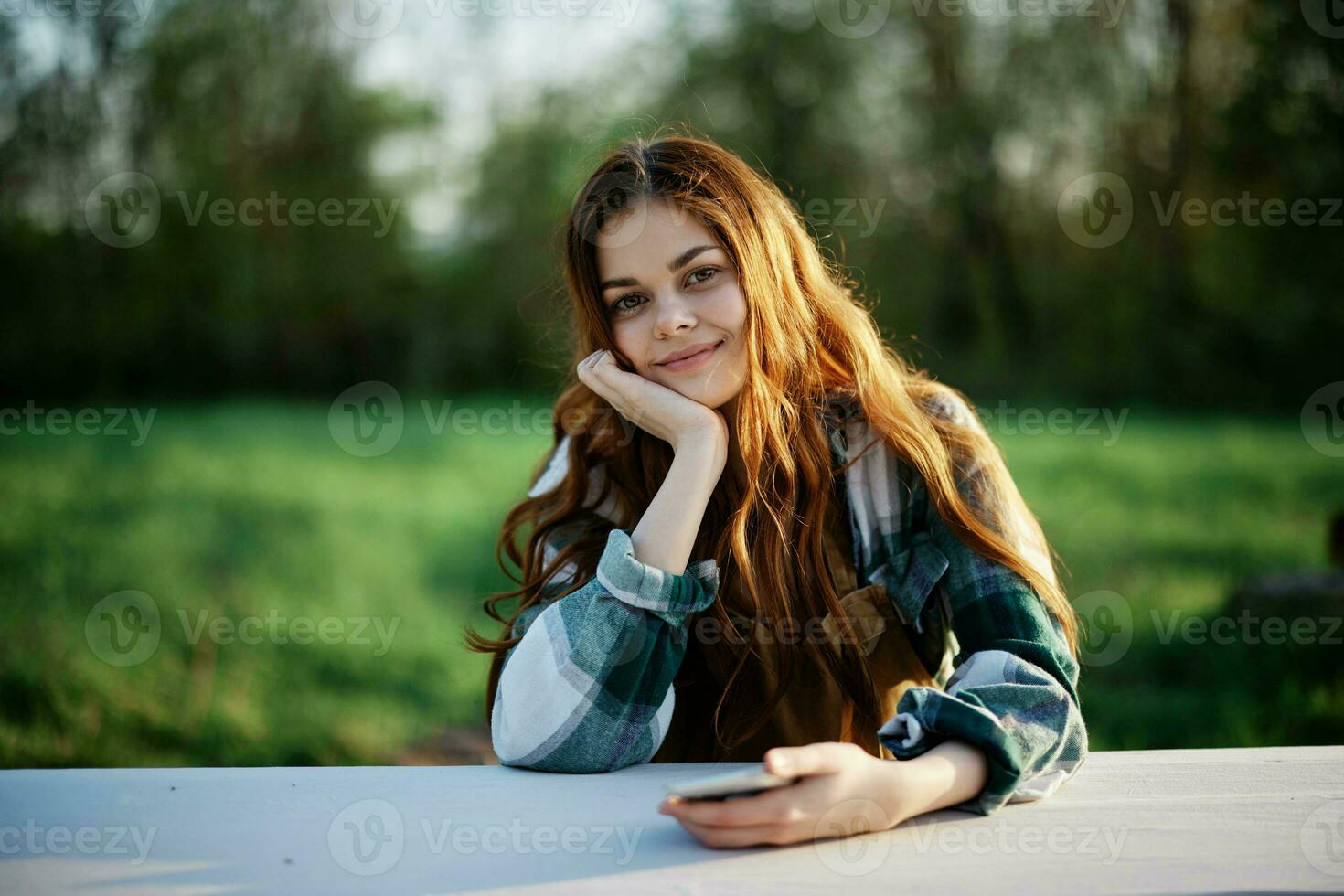 A beautiful woman relaxing and working on her phone sitting in nature in the park among the trees smiling and holding her smartphone in her hand lit by the bright sunset light photo