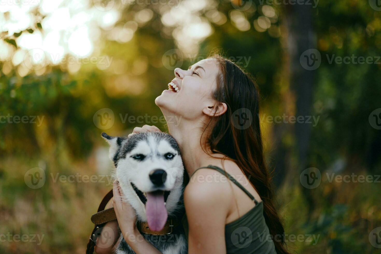 A woman with a husky breed dog smiles and affectionately strokes her beloved dog while walking in nature in the park in autumn against the backdrop of sunset photo