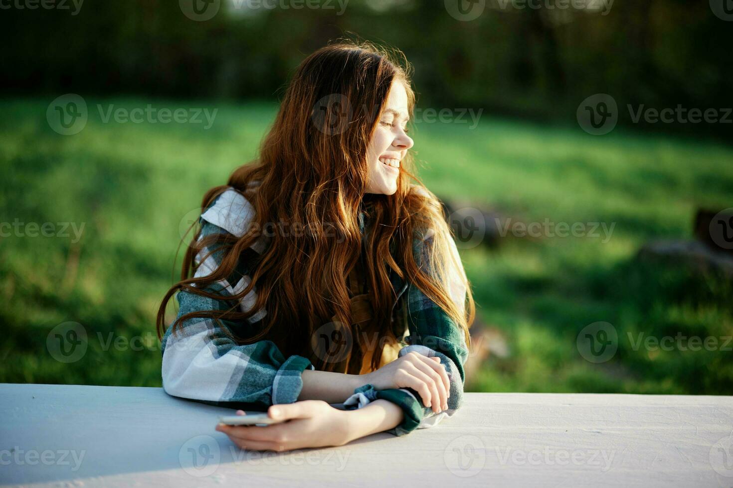 Girl with phone in hand sitting in nature in the park at the table smiling beautifully and looking at the camera with her red hair lit by the sunset sunlight of summer photo