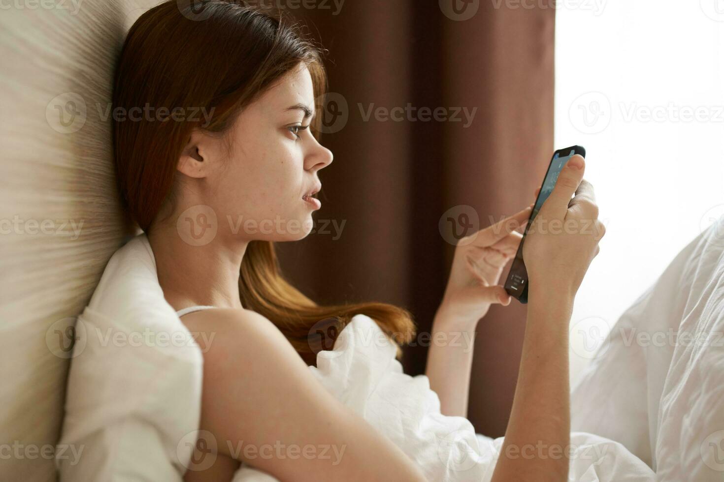 a woman under the covers lies in bed and looks at the mobile phone screen side view photo