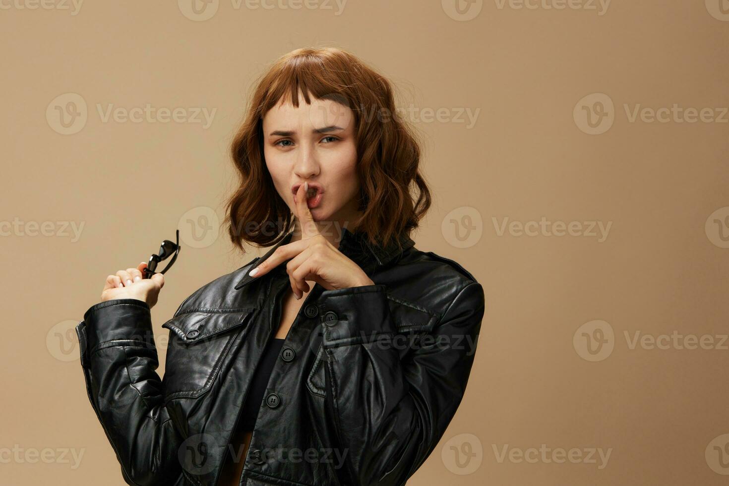 Happy stylish pretty redhead lady in leather jacket hold sunglasses posing isolated on pastel beige studio background. Copy space Banner Offer. Over black concept. Fashion Cinema Eyewear Ad photo