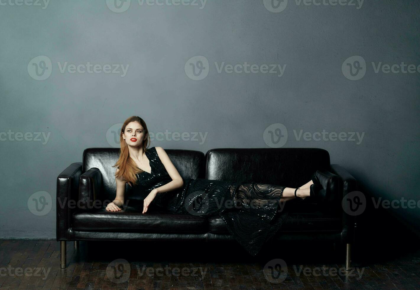 An elegant lady in a black dress lies on a leather sofa indoors in full growth photo