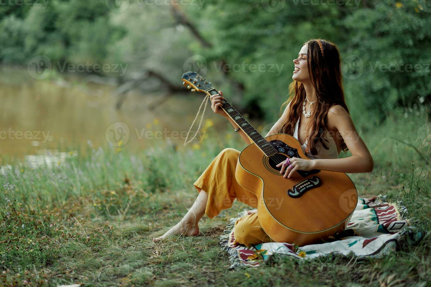 A hippie woman playing her guitar smiles and sings songs in nature sitting on a plaid in the evening in the sunset sunlight. A lifestyle in harmony with the body and nature photo