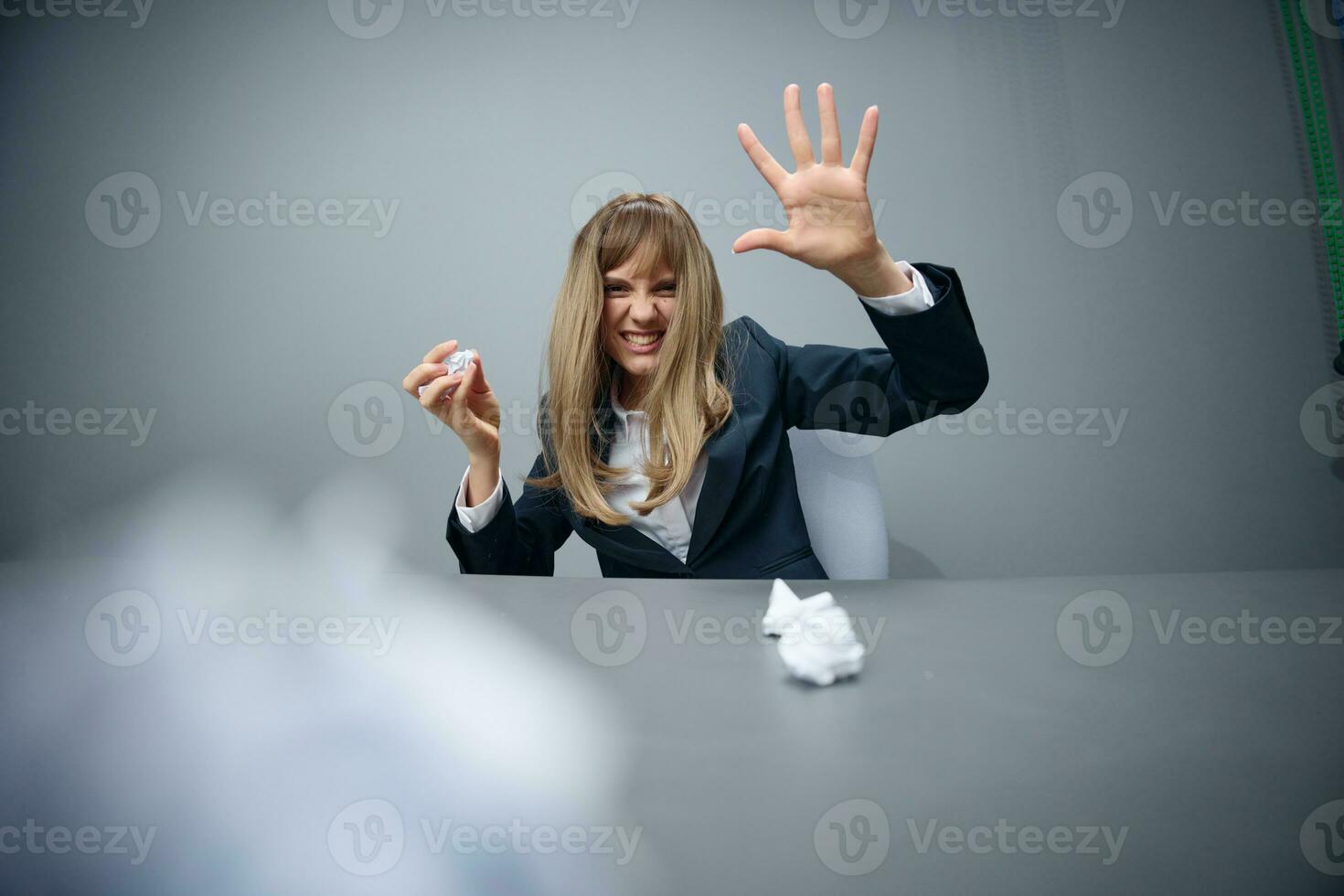 Funny millennial blonde businesswoman worker in blue jacket throws papers into the camera sitting at workplace in gray modern office. Tired of work Concept. Copy space, wide angle photo