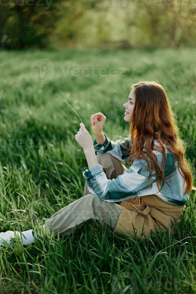 A woman sits on the green grass in a park with her back to the camera and relaxes in nature in the summer sunset evening light photo
