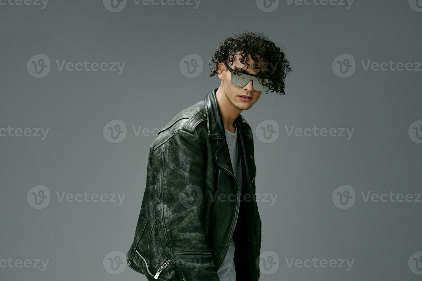 Stylish tanned curly man leather jacket trendy mirror sunglasses posing isolated on over gray studio background. Cool fashion offer. Huge Seasonal Sale New Collection concept. Copy space for ad photo