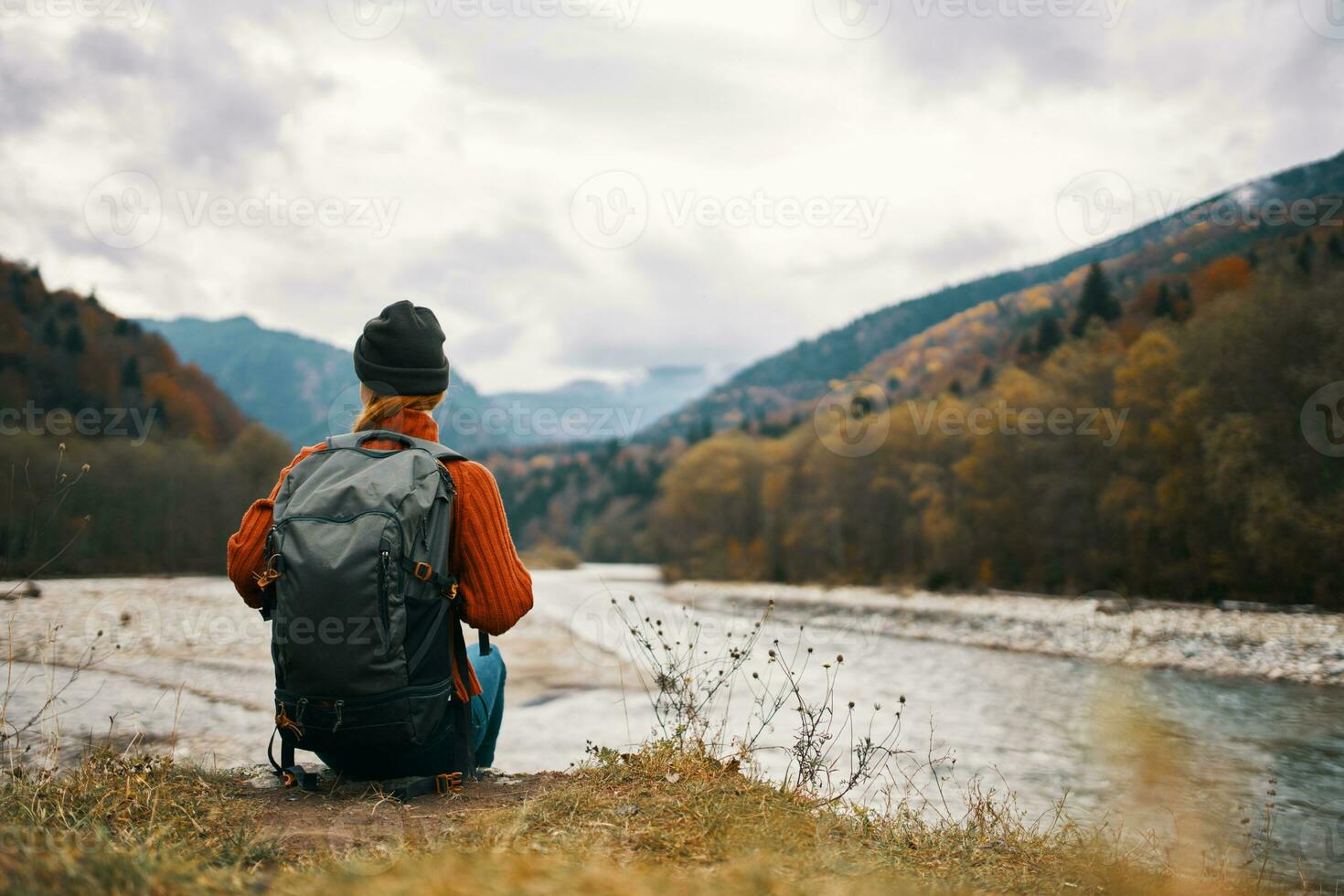 woman with a backpack and in a jacket with a hat on the river bank in the mountains in nature photo