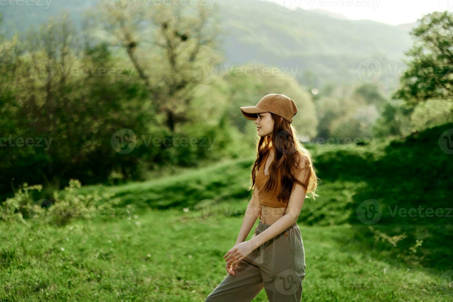 A beautiful woman in sportswear and a cap walks against a green natural landscape and smiles in the sunlight photo