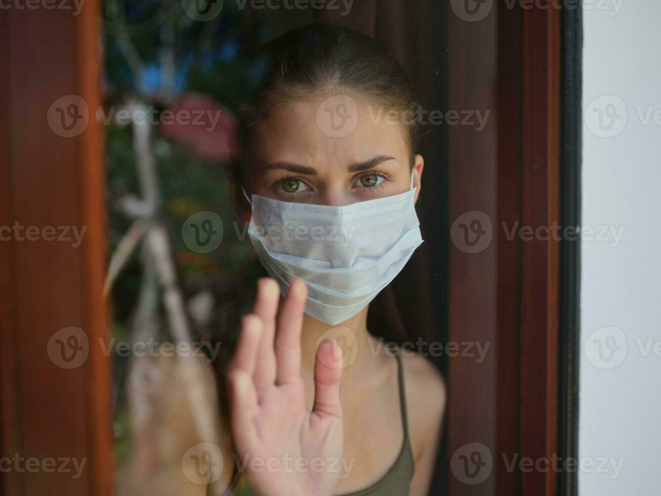 woman with a sad look in a medical mask looks out the window photo