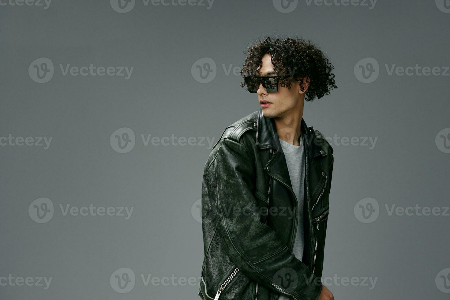 Stylish tanned curly man leather jacket in mirror trendy sunglasses posing isolated on over gray studio background. Cool fashion offer. Huge Seasonal Sale New Collection concept. Copy space for ad photo