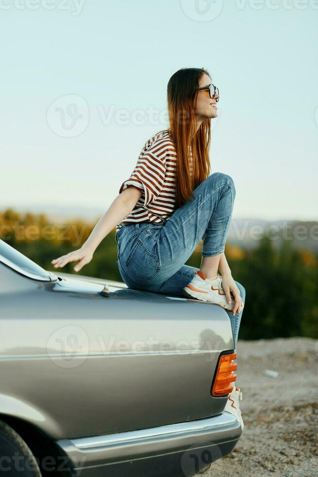 un Moda mujer en elegante anteojos, un a rayas camiseta y pantalones se sienta en el maletero de un coche y mira a el hermosa naturaleza de otoño foto