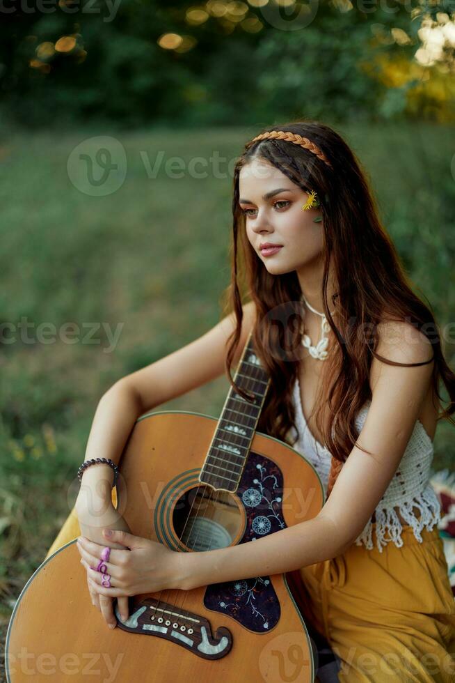 hippie mujer eco simpático jugando guitarra sonrisas y canto canciones en naturaleza sentado en un tartán por el lago en el noche en el puesta de sol luz de sol. un estilo de vida en armonía con el cuerpo y naturaleza foto