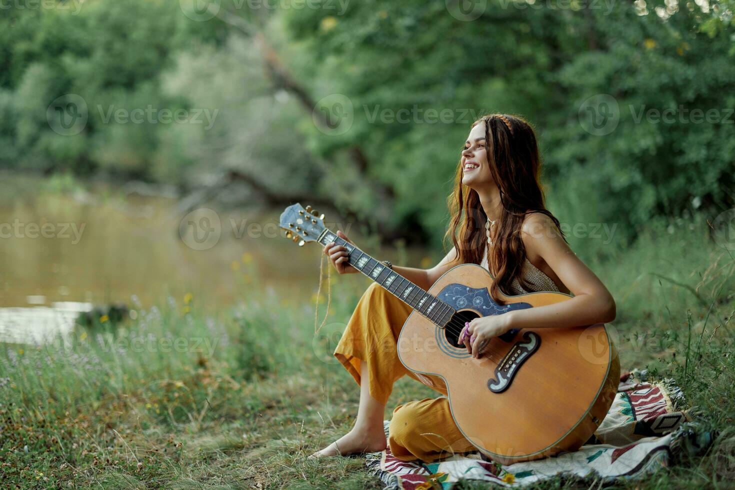 Young hippie woman sitting outdoors on a plaid and playing her guitar on  the riverbank into the sunset 23688263 Stock Photo at Vecteezy