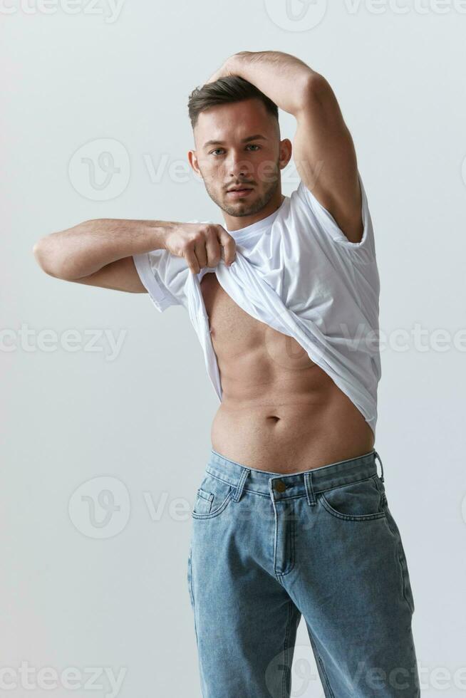 Portrait shot of handsome sexy flirting tanned man guy raising basic t-shirt looks at camera posing on white background. Fashion Style New Collection Offer. Copy space for ad. Model snap photo