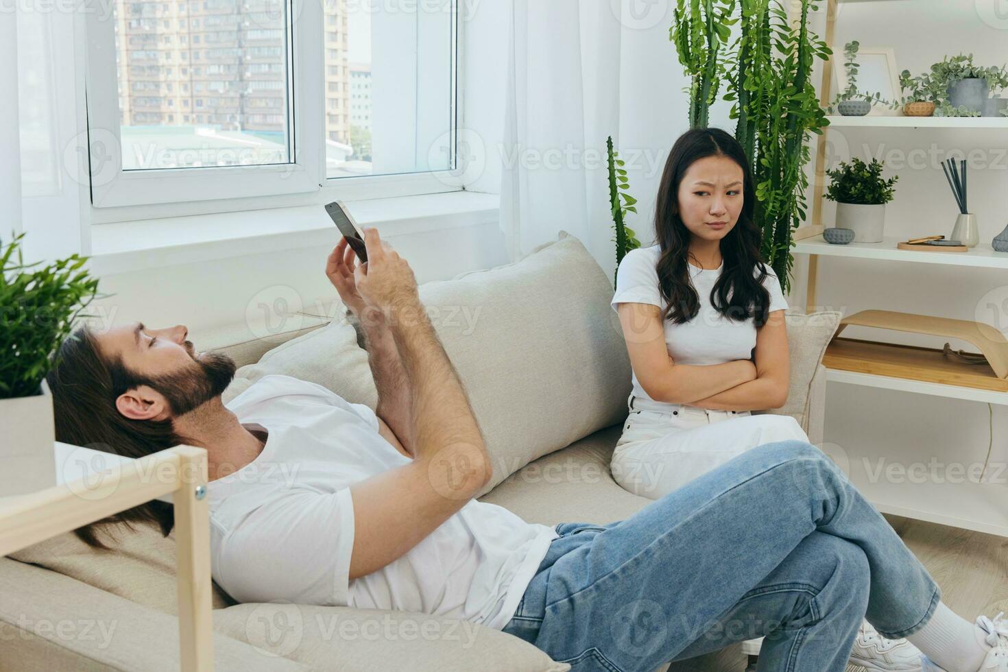 A man looks at his phone screen and smiles during an argument with his girlfriend. The angry and hurt woman looks in his direction and is sad. Family discord at home, phone addiction photo