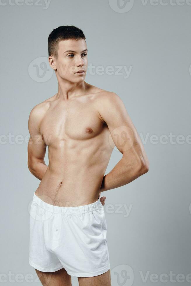 un hombre en blanco pantalones cortos sostiene su manos detrás su espalda un Mira a el lado inflado torso rutina de ejercicio foto