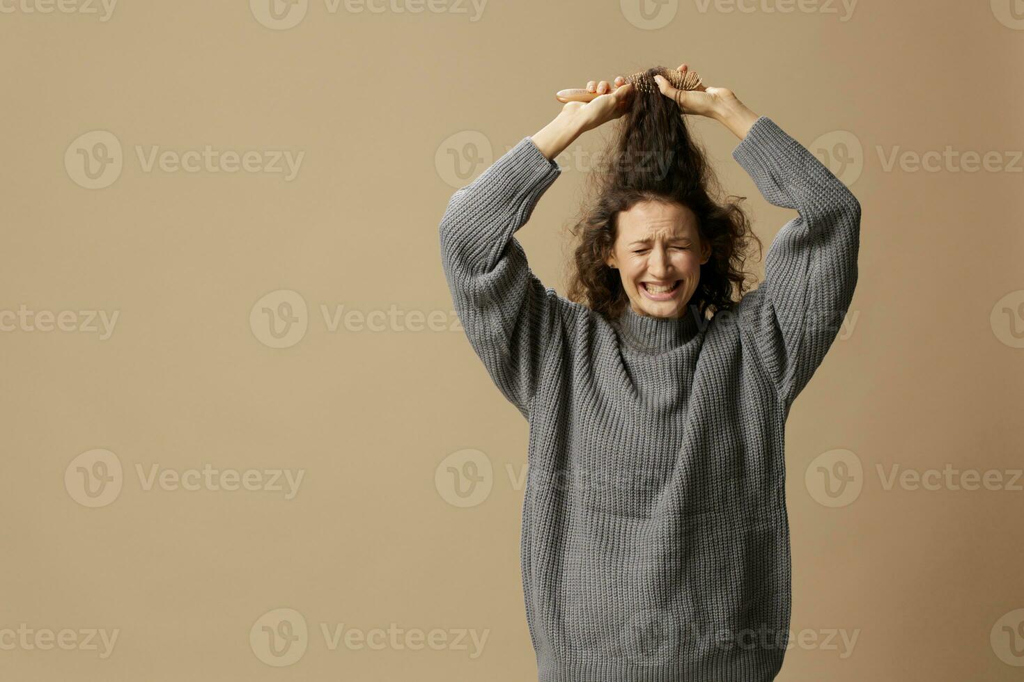 Problematic unruly damaged hair concept. Suffering curly beautiful woman in gray casual sweater with hairbrush comb pulls tangled hair up posing isolated on over beige pastel background. Copy space photo
