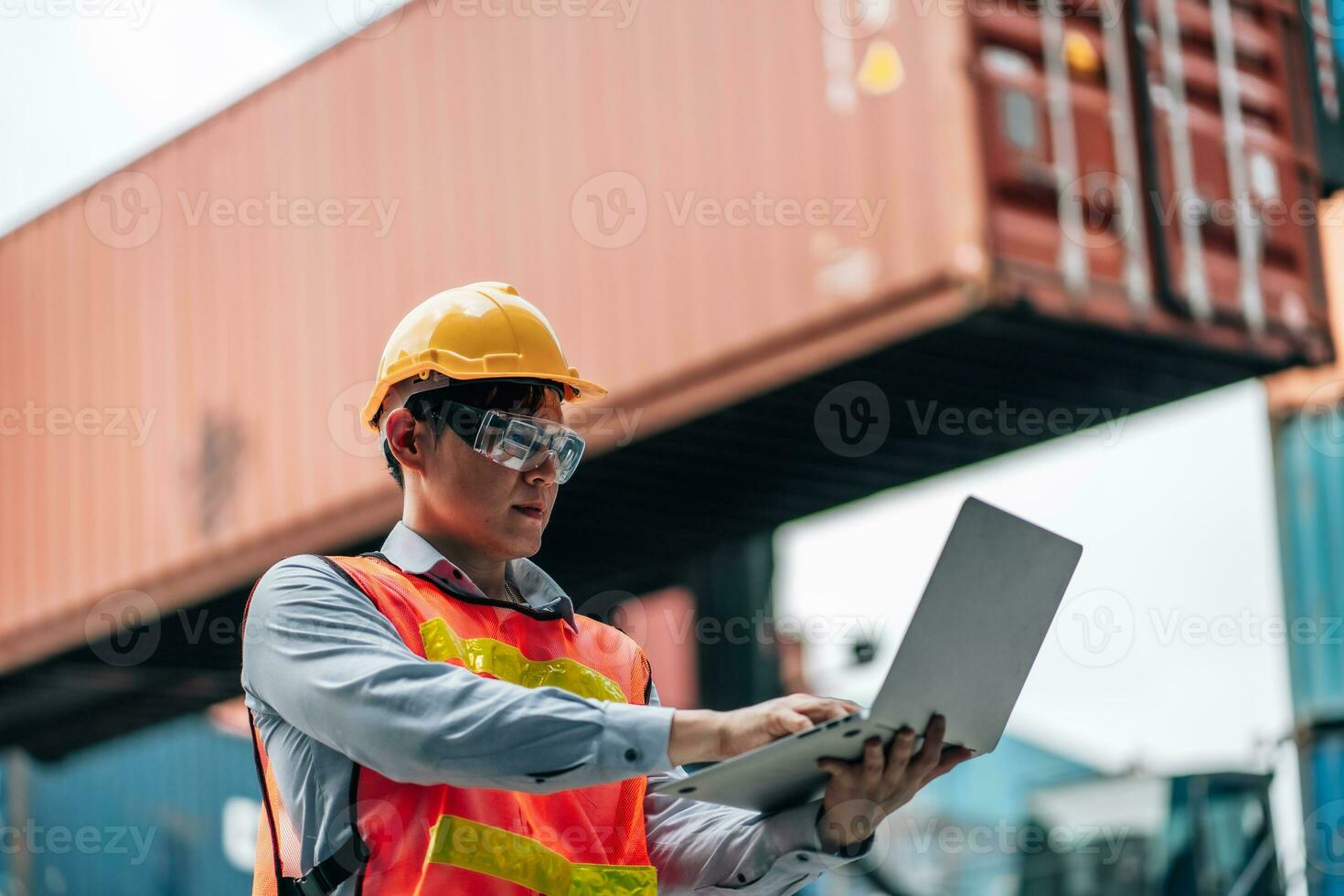 Asian technician dock worker in protective safety jumpsuit uniform and with hardhat and use laptop computer at cargo container shipping warehouse. transportation import,export logistic industrial photo
