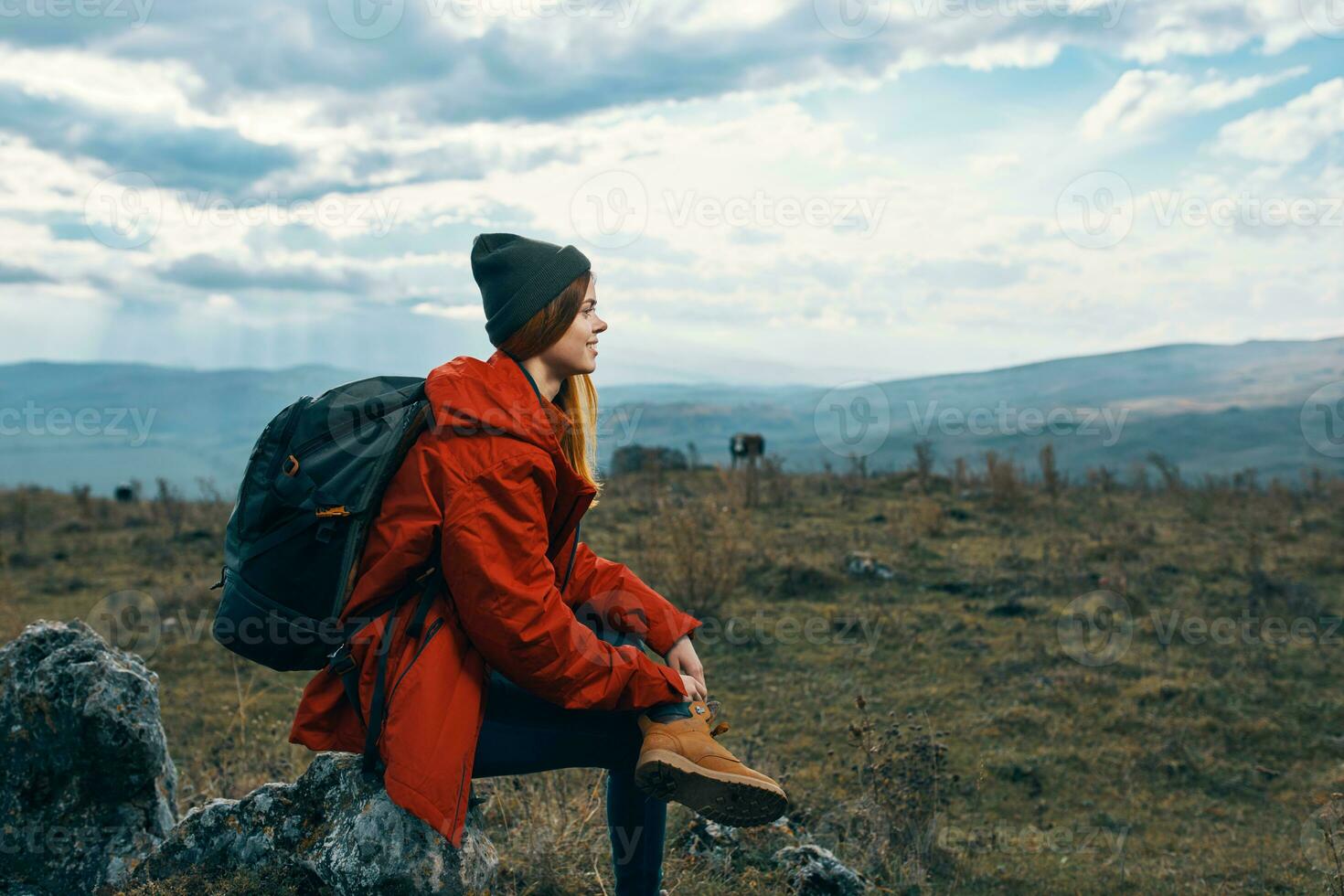 Traveler In a red jacket, hats with a backpack sits on a stone in the mountains in nature photo
