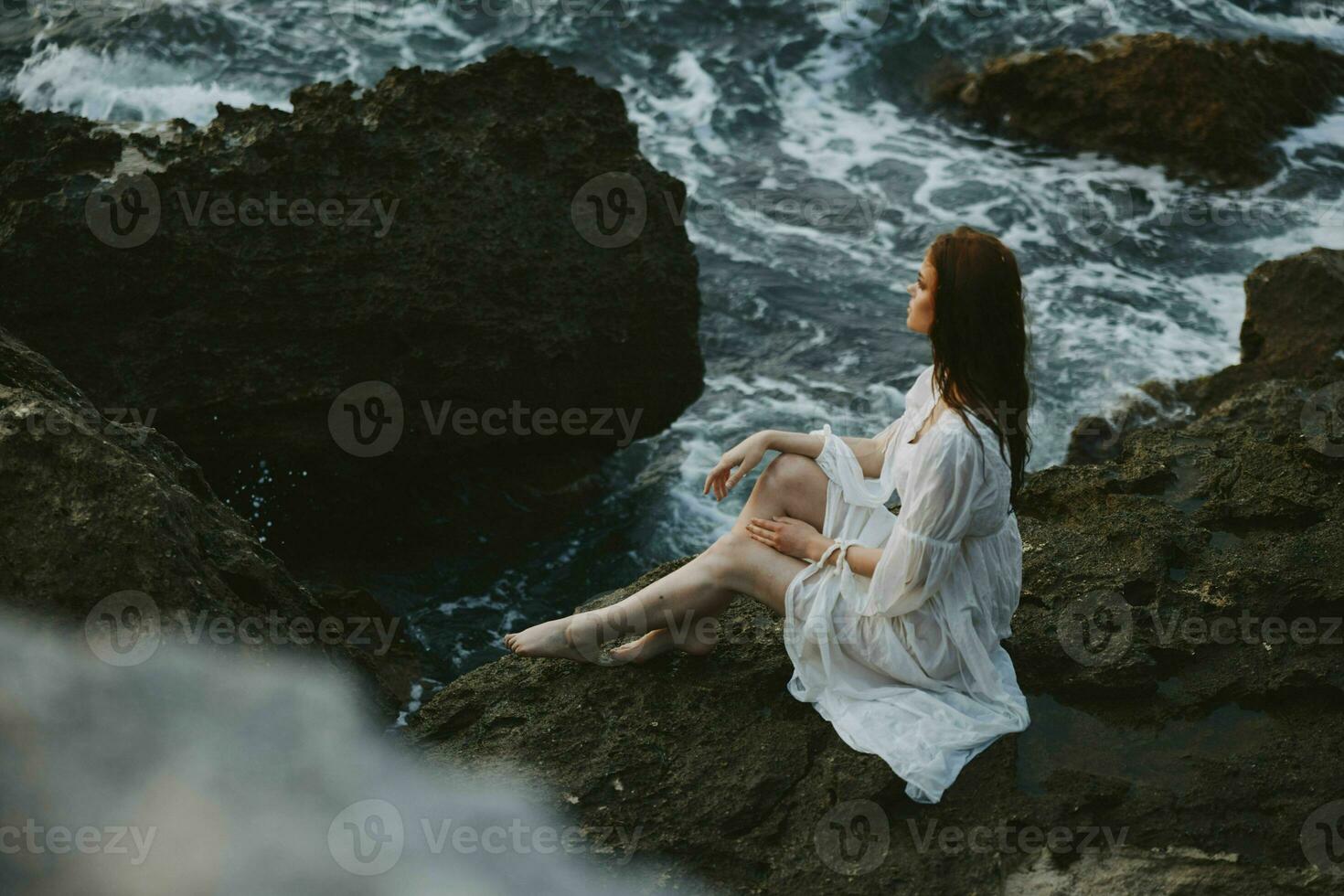 Barefoot Woman in White Dress with Wet Hair Sits on a Cliff photo