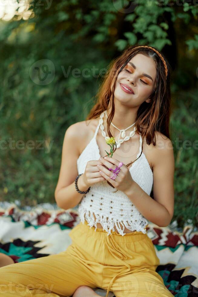 Young hippie woman sitting in nature on a plaid with a flower in her hands and looking at the camera smiling sincerely in eco-clothing in autumn in the sunset photo