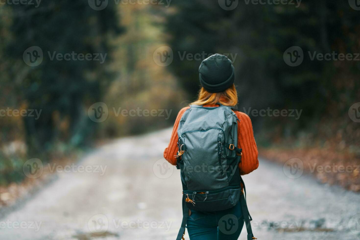 woman with a backpack in a hat and an orange sweater on the road in the autumn forest back view photo