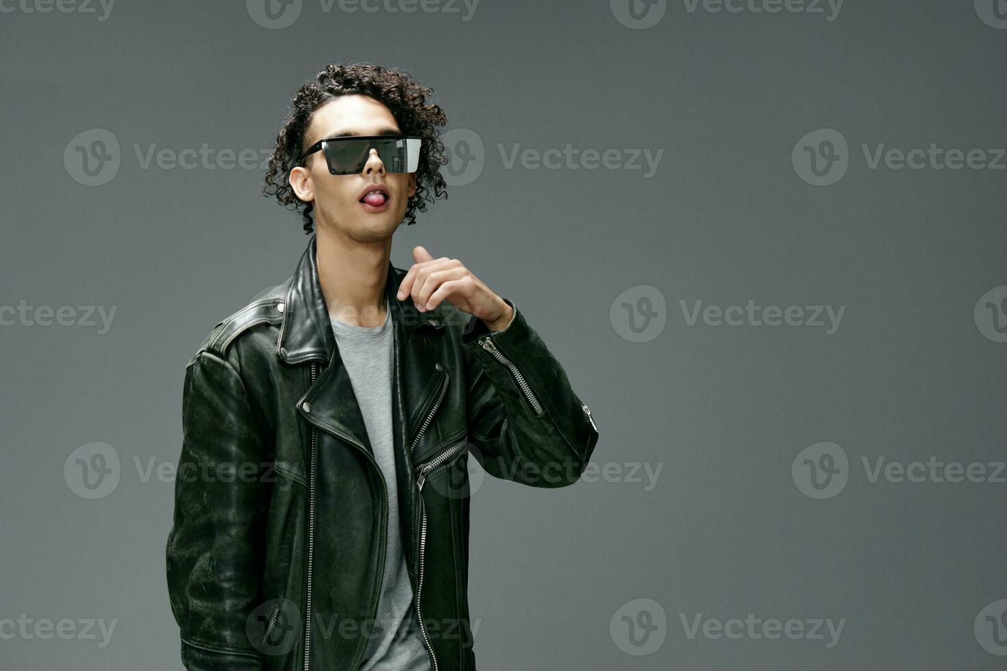 Side of view of stylish tanned curly man leather jacket trendy sunglasses posing isolated on gray studio background. Cool fashion offer. Huge Seasonal Sale New Collection concept. Copy space for ad photo