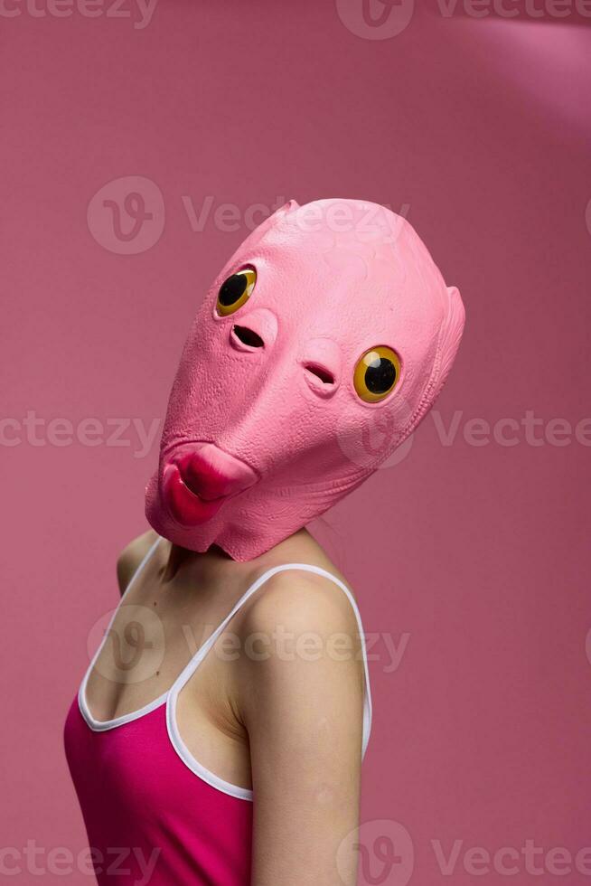 Woman wearing a pink fish head Halloween mask stands and looks at the camera against a pink background photo