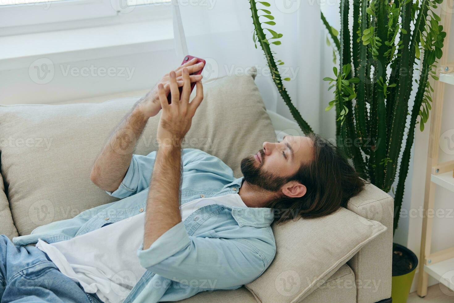 A man with a beard lies on the couch during the day at home and looks at his phone relaxing on his day off, a man gambling on the stock market online on his phone photo